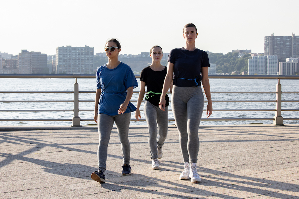 Three dancers sternly walking forward in front of a city scape. one wears sunglasses all wear t-shirts and leggings 